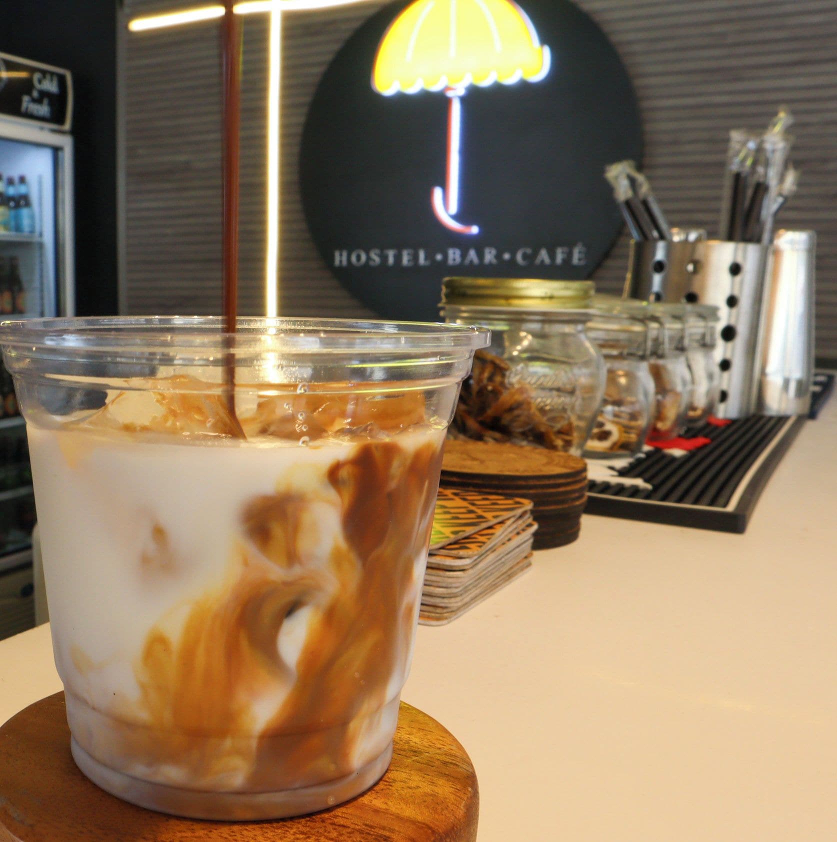 A clear plastic cup with a layered beverage, possibly iced coffee, on a wooden coaster. In the background, there is a sign for a hostel, bar, and café.