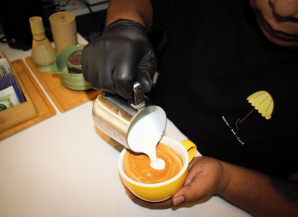 A person pouring a liquid into a cup, wearing a black apron with a logo, and holding a yellow cup.