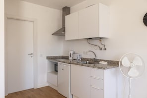 Une cuisine compacte avec des meubles blancs, un plan de travail en granit, un lave-vaisselle encastré, une machine à café, un évier avec un robinet et un ventilateur fixé au mur.