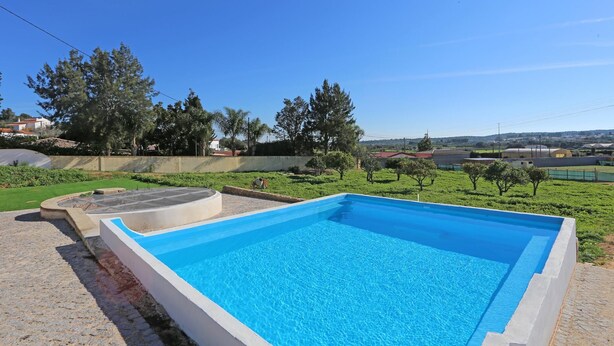 A rectangular swimming pool with a clear blue water surface, surrounded by a low stone wall and a grassy area with trees. - X 06 - 1 Bed Apartment in Quinta da Ponte (Lagos)