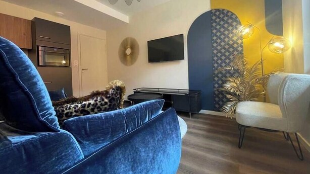 A modern living room with a blue sofa, a flat-screen TV, a ceiling fan, and a patterned wall. - Suite "Art Déco" - 2/4 Pers (Niergnies)