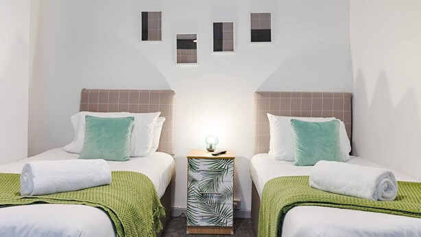 Une chambre d’hôtel avec deux lits, une table de chevet avec une lampe et une petite étagère fixée au mur, ornée d’objets décoratifs.