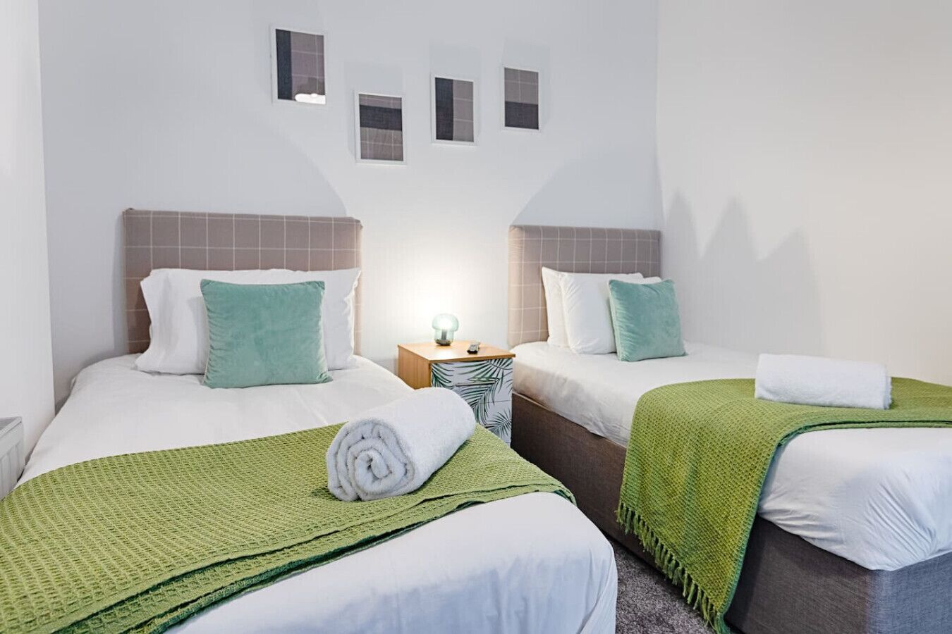 A hotel room with two beds, each with a green blanket, white pillows, and a small bedside table with a lamp.
