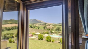 A window with a view of a grassy landscape, a swimming pool, and a hill in the distance. - A cosy farmhouse surrounded by nature! (Marche)