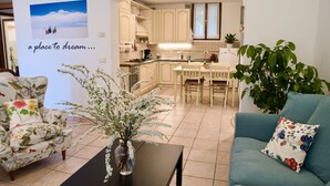 A living room with a sofa, a chair, a coffee table, and a vase with flowers. There is a kitchen in the background with cabinets and a window. - Happy Corner - The Home for Outdoor Lovers - (Veneto)