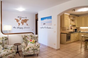 Uma sala de estar com poltronas estampadas, uma mesa redonda pequena, um mapa-múndi como decoração na parede e uma cozinha com armários em tons claros.