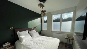 Un dormitorio con una cama grande, un ventanal con vista a un lago, un televisor montado y una mesita pequeña.