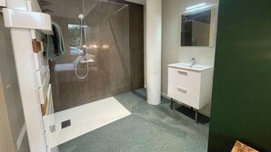 Un baño moderno con cabina de ducha de vidrio, un tocador blanco con espejo y un piso gris con textura.