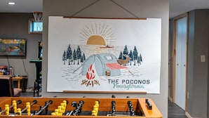 Une pièce équipée d’un baby-foot et ornée d’une œuvre d’art murale représentant une scène de camping avec une tente, des arbres, un feu de camp et un soleil.