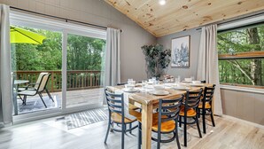 Un espace repas avec une table et des chaises en bois, de grandes fenêtres avec des rideaux et une vue sur la verdure à l’extérieur.