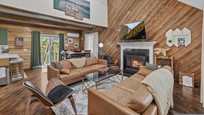 A modern living room with a fireplace, leather sofas, and a dining area with a view. - Dog Friendly Cabin w Hot Tub Sauna Fire Pit (Tobyhanna)