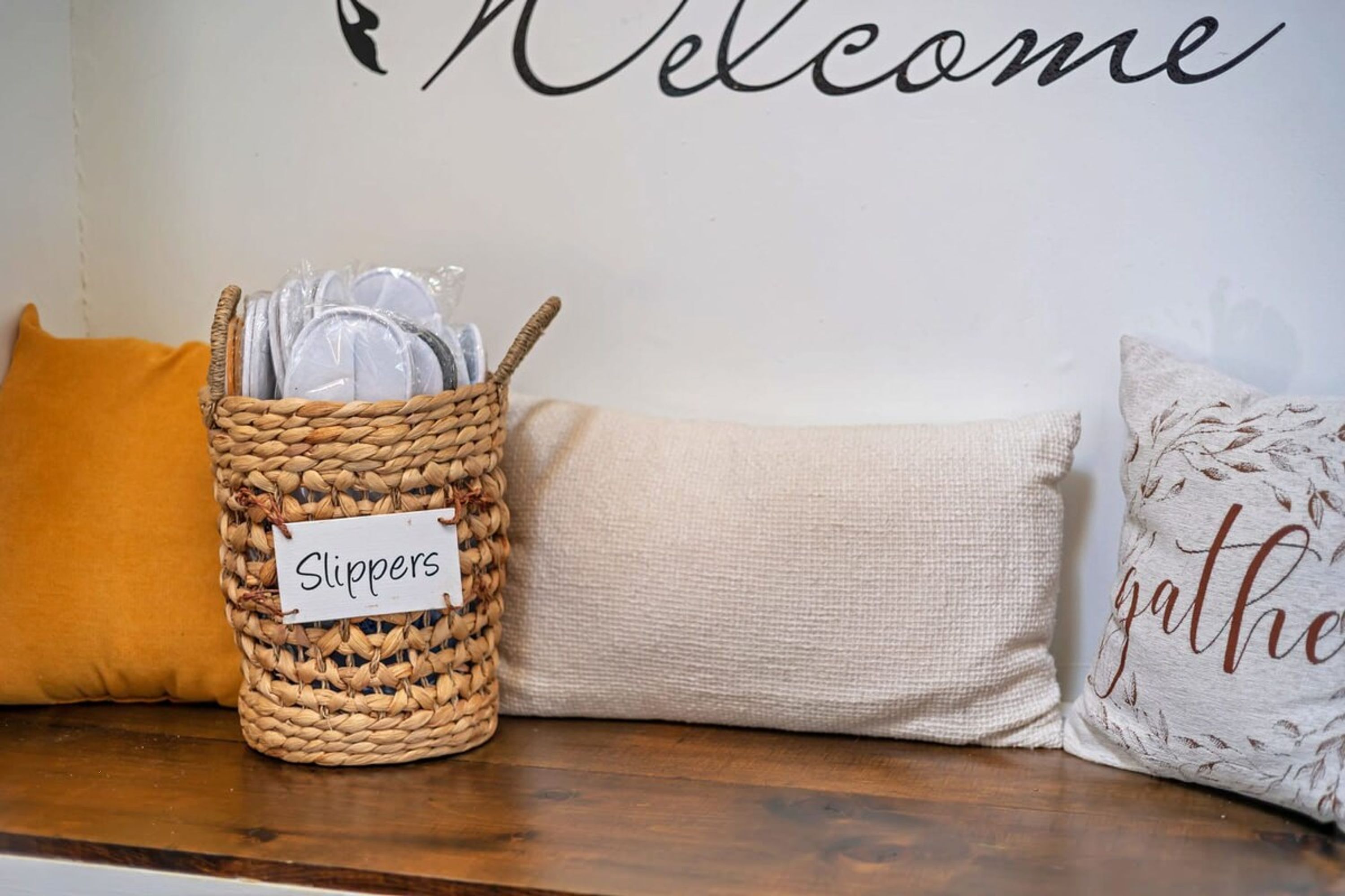 A wicker basket labeled 'Slippers' on a wooden surface, next to a beige cushion and a pillow with 'gather' text.