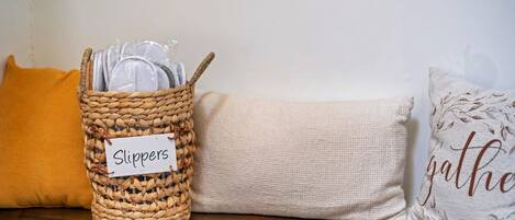 A wicker basket labeled 'Slippers' on a wooden surface, next to a beige cushion and a pillow with 'gather' text.