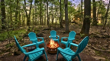 A forest with a fire pit and four turquoise chairs arranged around it.