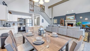A modern kitchen with stainless steel appliances, a dining area with a wooden table set for four, and a living room with a fireplace and a large painting. - Near Jfbb for Skiing With Hot Tub and Fire Pit (Blakeslee)