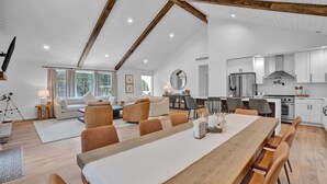 Uma sala de estar espaçosa com uma longa mesa de jantar de madeira, uma cozinha com eletrodomésticos modernos e um teto alto com vigas aparentes.