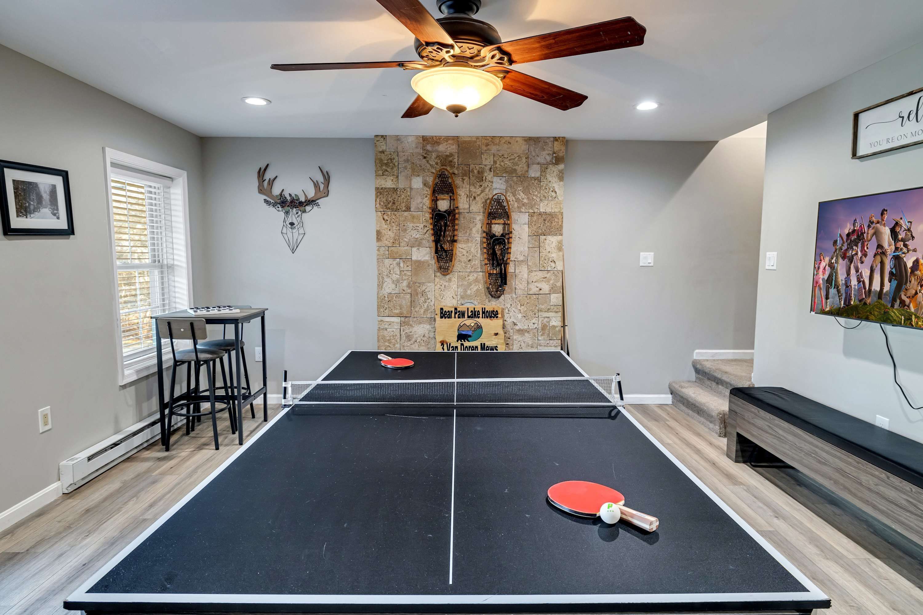 Une table de ping-pong dans une pièce avec un mur en pierre, un ventilateur de plafond et un tableau encadré accroché au mur.