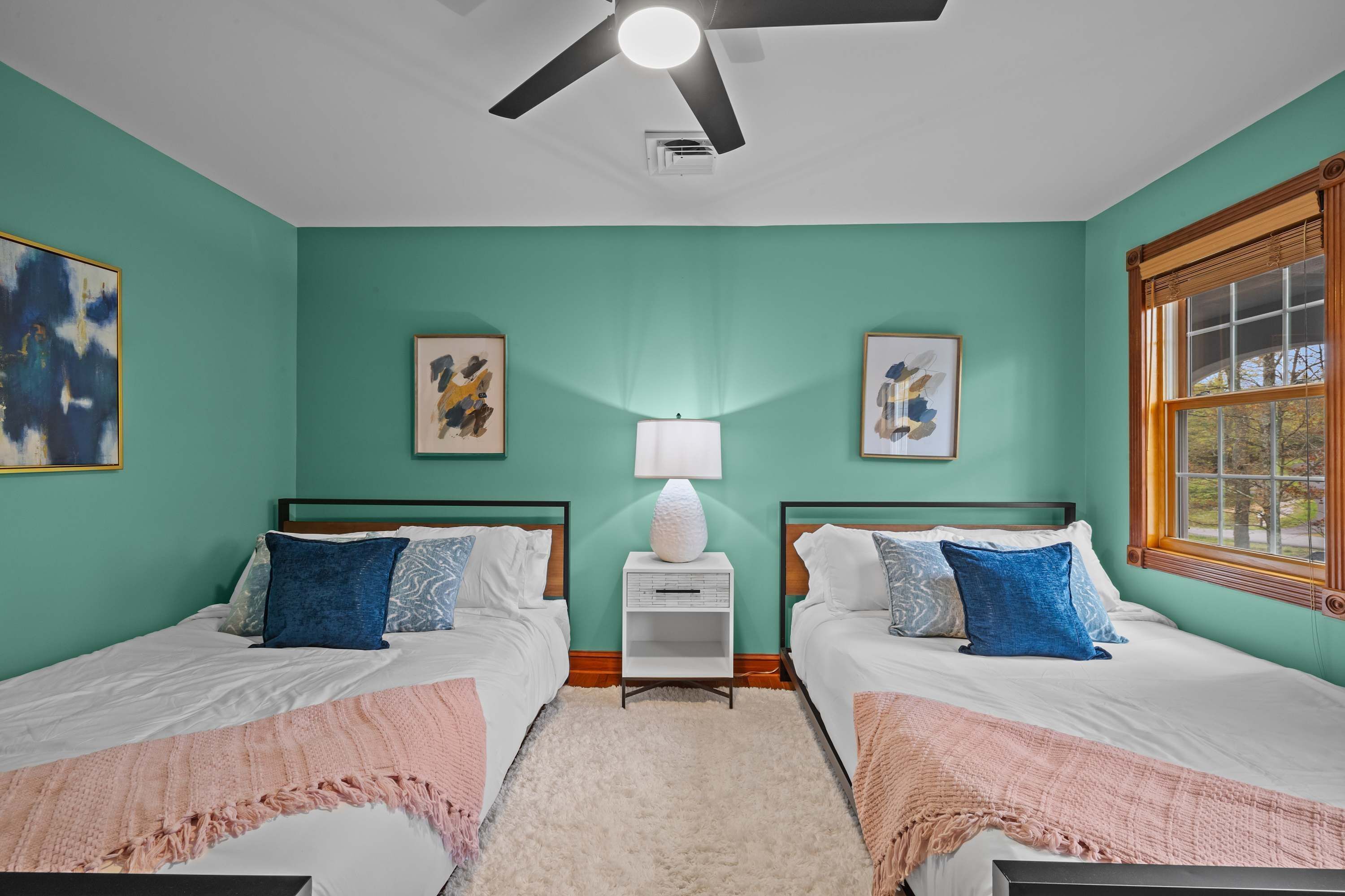 A bedroom with two beds, a ceiling fan, a window, and artwork on the walls.