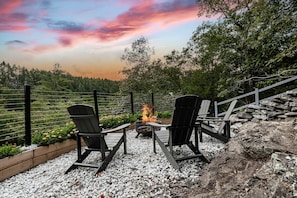 Een terras met Adirondack-stoelen, een vuurkorf en uitzicht op een bebost gebied.