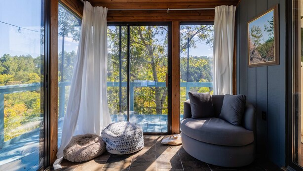 A room with a large sliding glass door, a gray sofa, a woven pouf, and a framed picture on the wall. - Riverfront Dog Friendly Cabin With Mountain Views (East Stroudsburg)