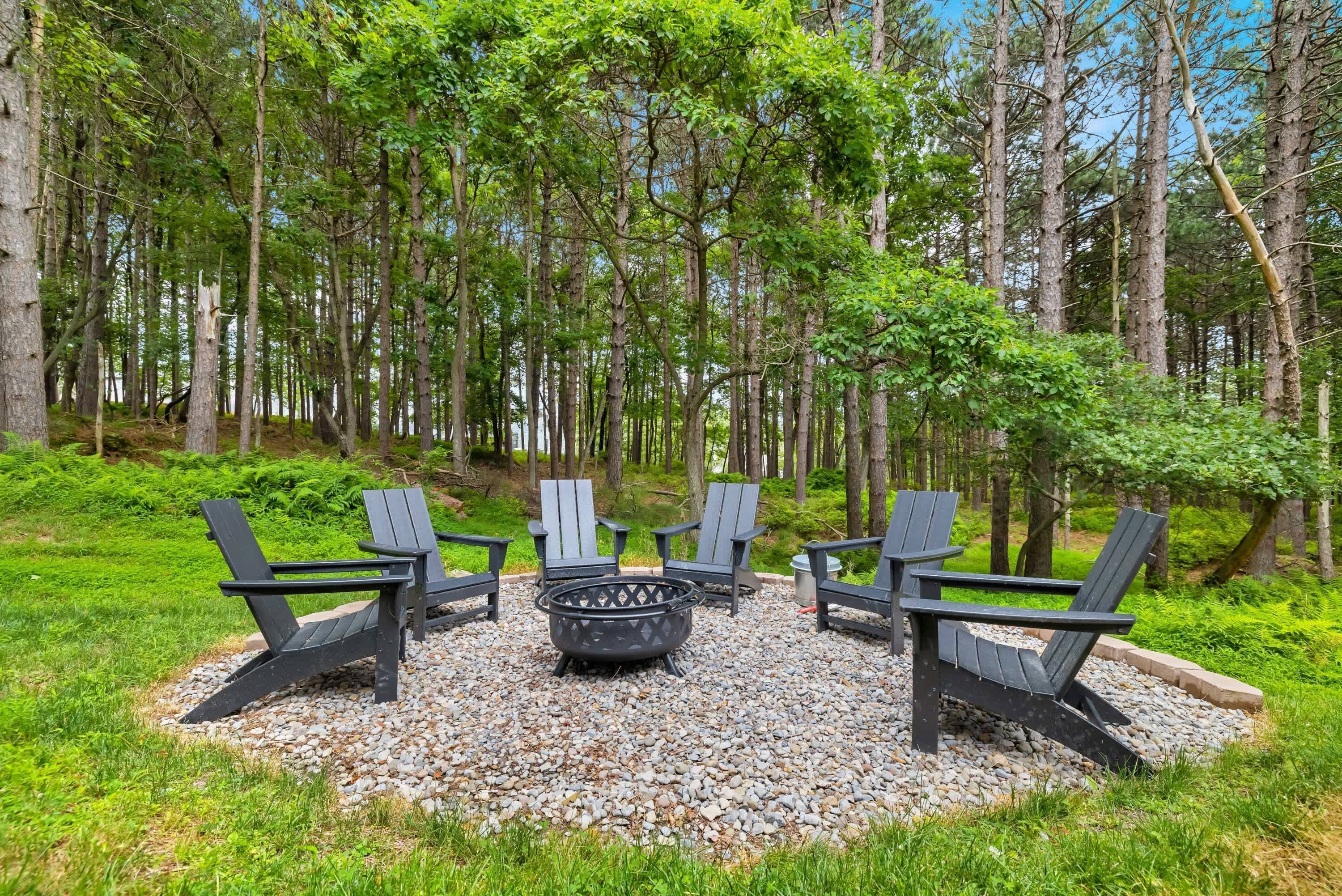 A forested area with a fire pit and four Adirondack chairs arranged in a circle.