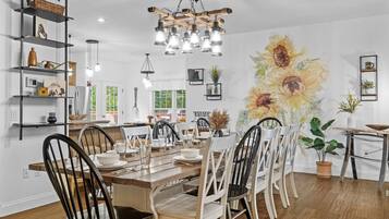 A dining area with a wooden table, white chairs, and a chandelier. There are shelves with various items and a large sunflower mural on the wall.