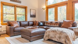 A living room with a sectional sofa, ottoman, and large windows offering a view of trees. - Whiteface Views Hot Tub Game Room Dog Friendly (Wilmington)
