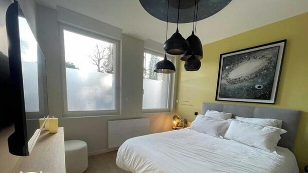 A bedroom with a bed, bedside table, two candles, a round stool, a window, and a wall-mounted artwork. - Suite "Petit Prince" - 2/4 Pers (Niergnies)