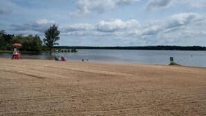 A sandy beach with a lifeguard tower, a red umbrella, and a calm lake under a cloudy sky. - Near Jfbb With Private Hot Tub and Game Room (Albrightsville)