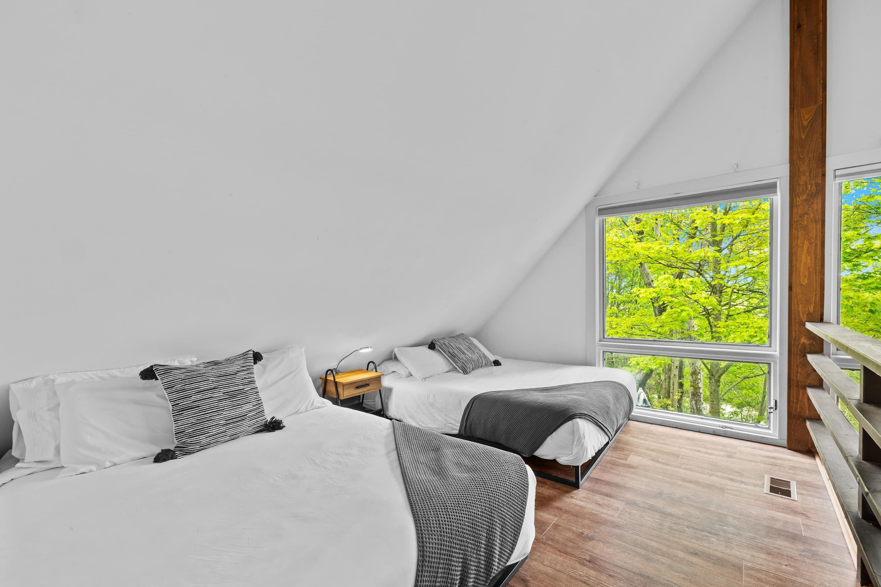Un dormitorio moderno con dos camas, piso de madera y un ventanal con vistas a árboles.