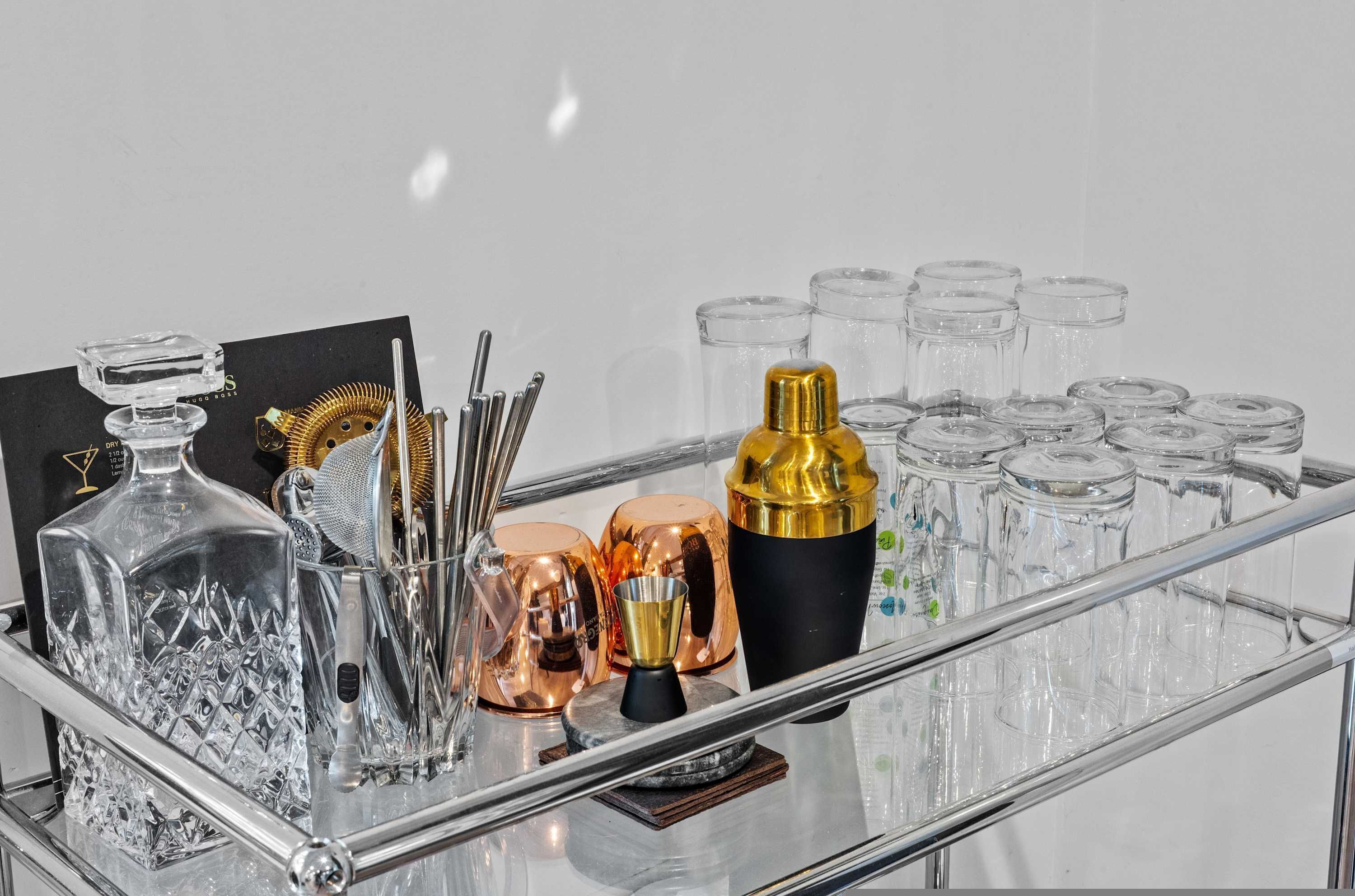 A bar setup with various glassware, a gold cocktail shaker, and a metal strainer on a glass shelf.