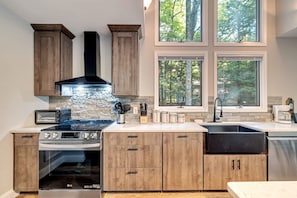 A modern kitchen with a stove, oven, and microwave, wooden cabinets, and a large window with a view of trees. - Dog Friendly Escape w Gameroom Hot Tub Fire Pit (Lake Harmony)