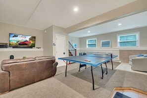 Una sala de estar con mesa de ping pong azul, un sofá de cuero marrón, un televisor mostrando un partido y una escalera que conduce a otro nivel.