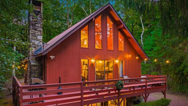 A cabin with a stone chimney, a deck with seating, and a large window overlooking the forest. - Poconos Speakeasy Hot Tub Dog-friendly Near Ski (Lake Harmony)