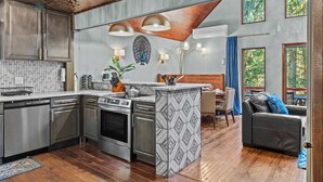 A modern kitchen with stainless steel appliances, a dining area with a wooden table and chairs, and a view of the outdoors through large windows. - Poconos Speakeasy Hot Tub Dog-friendly Near Ski (Lake Harmony)