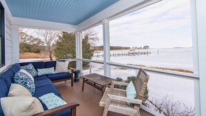 A covered porch with a blue ceiling, a blue sofa with patterned cushions, a wicker chair, and a view of a body of water. - Modern Chesapeake Waterfront - Heated Pool - Dock (Cambridge)