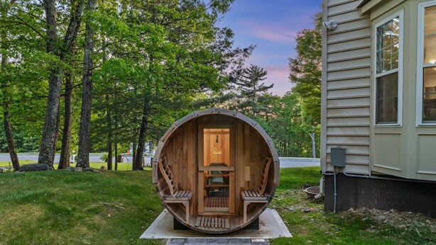 Eine Holzbottgeflechtkonstruktion mit einer Tür, Stufen und Bänken, gelegen in einer grünen Wiese mit Bäumen und einem Haus im Hintergrund.