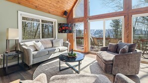 A cozy living room with a grey sofa, a round coffee table, and a grey armchair. A flat-screen TV is mounted on the wall, and a large window offers a view of trees and mountains. - Views of Blue Mountain Near App Trail Game Room (Palmerton)
