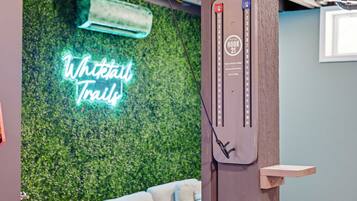 A room with a neon sign reading 'Whitetail Trails,' a wooden wall, a small wooden shelf, and a wall-mounted device.