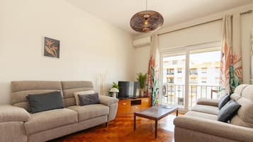Een woonkamer met een bank, een salontafel, een televisie op een houten standaard, en een balkon met uitzicht op gebouwen.