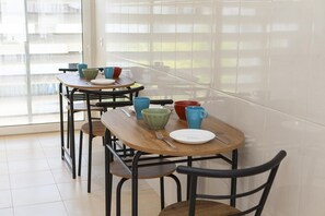 Uma sala de jantar com várias mesas e cadeiras, uma janela grande com persianas e vista para edifícios.