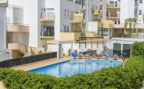 Piscina rodeada por espreguiçadeiras e um edifício com varandas.