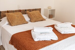 Un lit bien fait, agrémenté de deux coussins marron tissés, d’une couvre-lit blanche et d’un plaid marron. On y trouve également trois serviettes blanches pliées, chacune portant une inscription.
