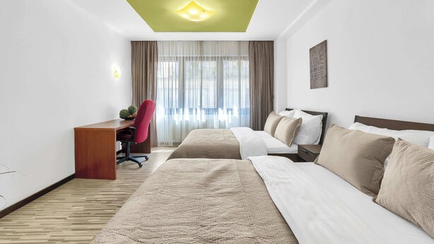 Habitación de hotel con escritorio, dos camas, una silla y un ventanal con cortinas.