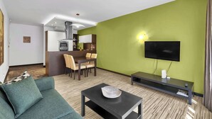 Una sala de estar con sofá, una mesa de centro, televisión, un comedor con sillas y una cocina al fondo.
