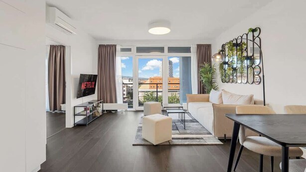 A modern living room with a flat-screen TV, a sofa, a dining table, and a balcony with a view of buildings. - Urban White Oasis in Centre (Bratislava - Old Town)