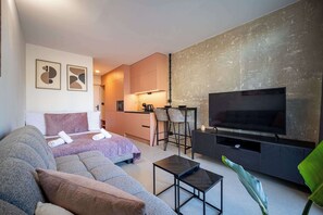 A modern living room with a sofa, a TV, a small coffee table, and a kitchen area in the background.