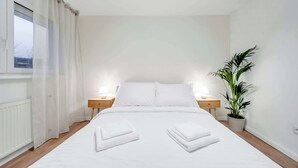 Un letto rifatto con due asciugamani piegati, un comodino con una lampada, una pianta in vaso e una finestra con tende leggere.