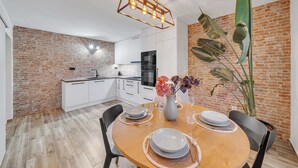 Una cucina moderna con mobili bianchi, un muro in mattoni e un'area pranzo con un tavolo di legno apparecchiato per quattro persone.