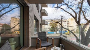 Un balcón con una silla, una mesa pequeña con una taza, y vistas a edificios y árboles.